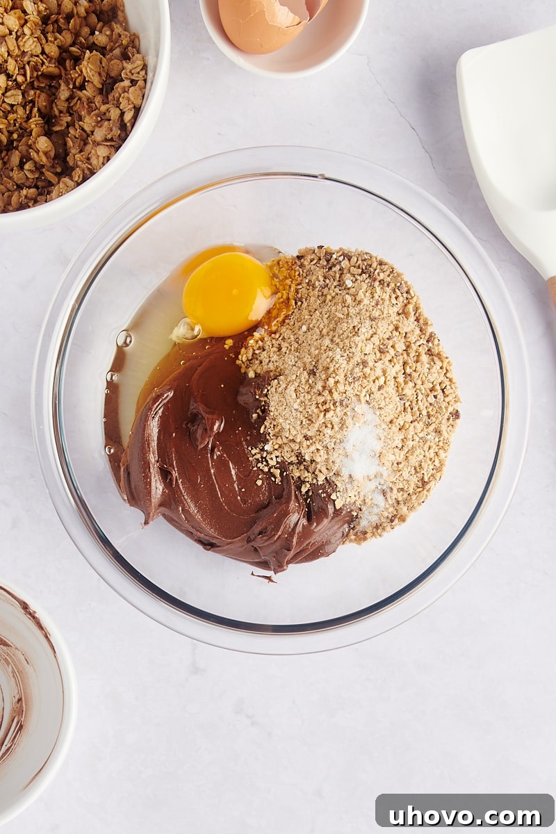 Ground granola, chocolate peanut butter, and a cracked egg combined in a mixing bowl, ready to be blended into cookie dough.