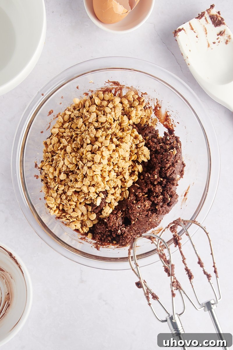 Whole granola pieces being added to the prepared cookie dough in a mixing bowl.
