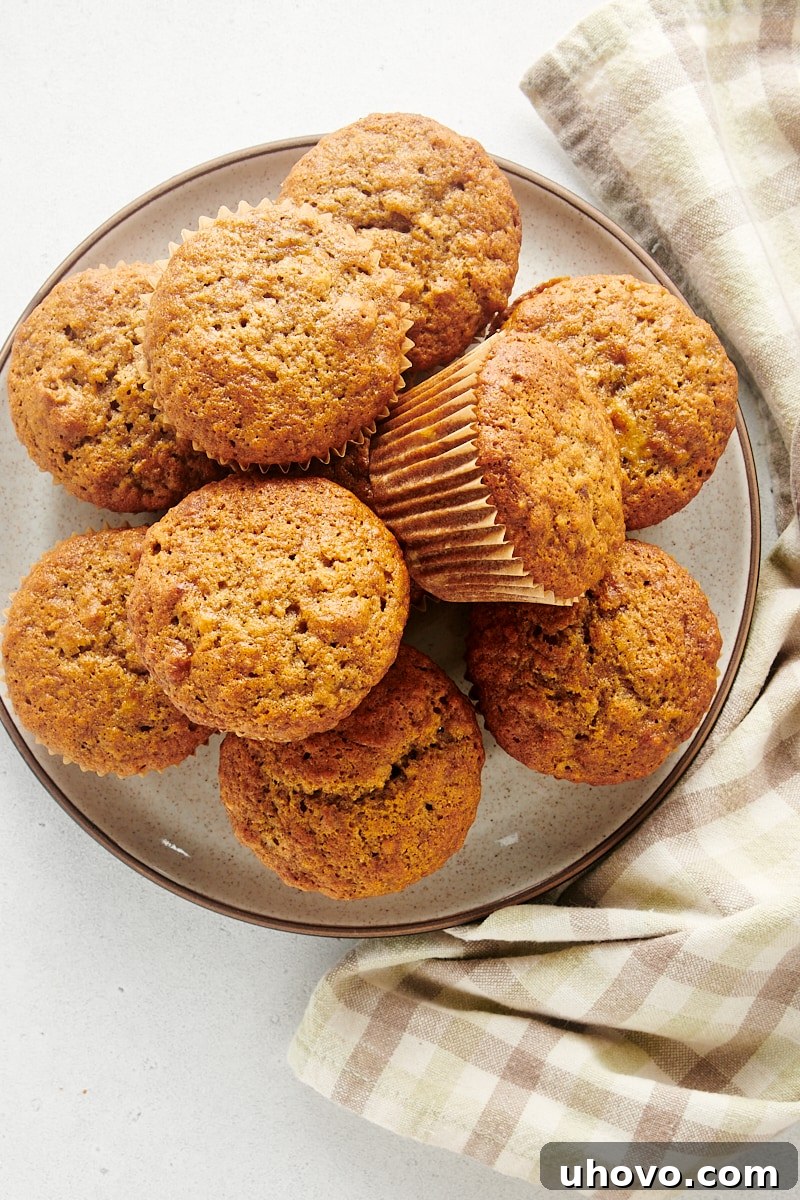Overhead view of a generous pile of freshly baked banana nut muffins, some with visible nut pieces, arranged on a white and brown speckled ceramic plate.