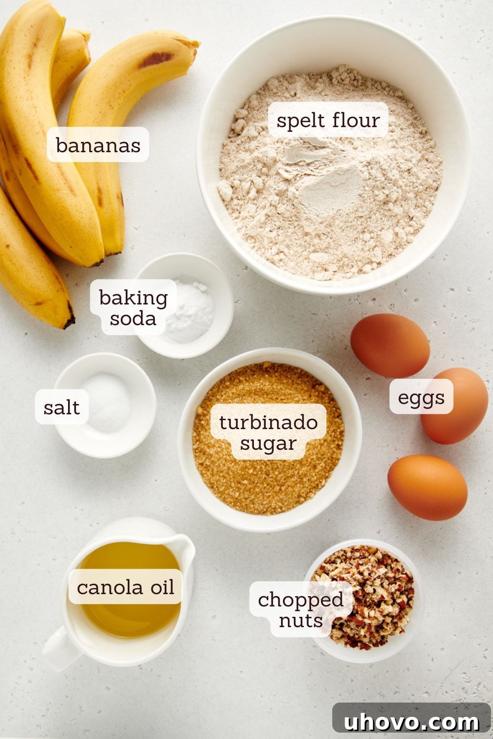 Overhead view of various ingredients for banana nut muffins laid out on a clean white surface, showcasing ripe bananas, eggs, flour, sugar, and nuts.
