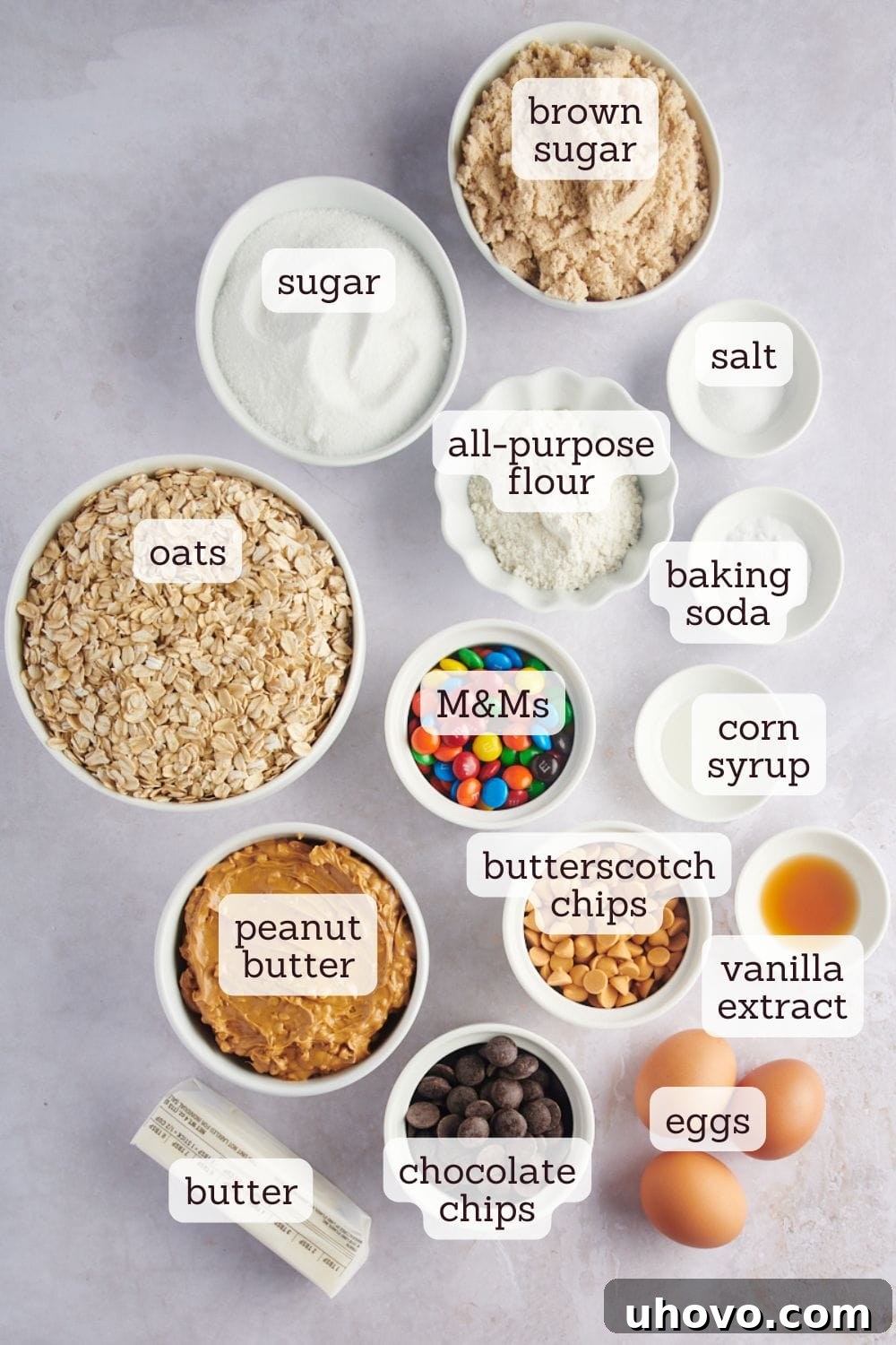 Overhead view of various ingredients for monster cookies laid out on a surface, including M&M's, chocolate chips, oats, and peanut butter.