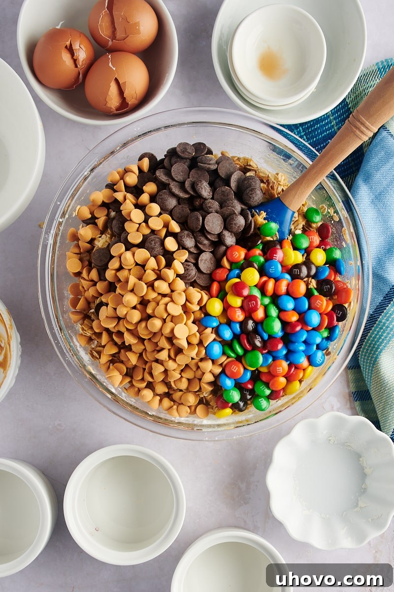 Mix-ins, including M&M's, chocolate chips, and butterscotch chips, folded into a bowl of monster cookie dough before chilling.