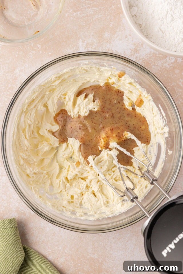 A step-by-step image showing chilled browned butter being added to a creamy mixture of beaten cream cheese and butter in a stand mixer, ready to be incorporated.