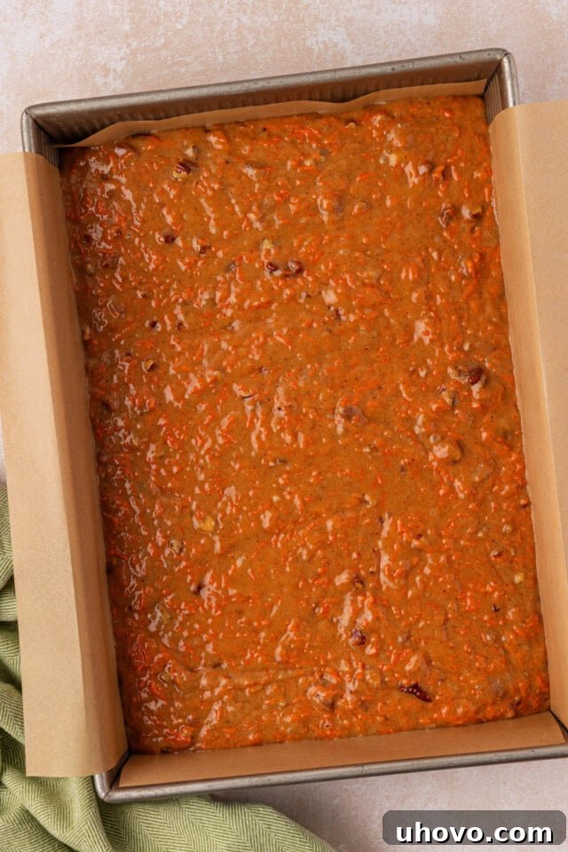 An overhead shot depicting carrot cake batter smoothly spread into a parchment-lined 9x13-inch baking pan, ready for the oven.