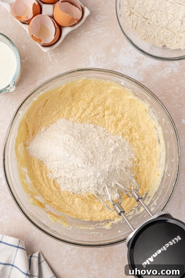 Overhead view of dry ingredients being added in portions to the creamy pound cake batter in a mixing bowl.