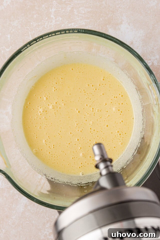 An overhead view of beaten eggs and granulated sugar, transformed into a pale, frothy, and slightly thickened mixture in the bowl of a stand mixer, indicating proper aeration.