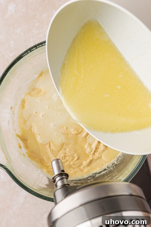 An overhead shot of the hot milk mixture being slowly and steadily poured into the cake batter in a stand mixer, creating a smooth, well-combined consistency.