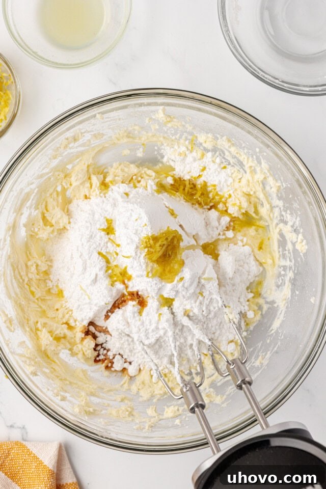 Confectioners' sugar, vanilla, lemon juice, and lemon zest being added to a bowl of perfectly beaten cream cheese and butter, ready to be whipped into the filling.