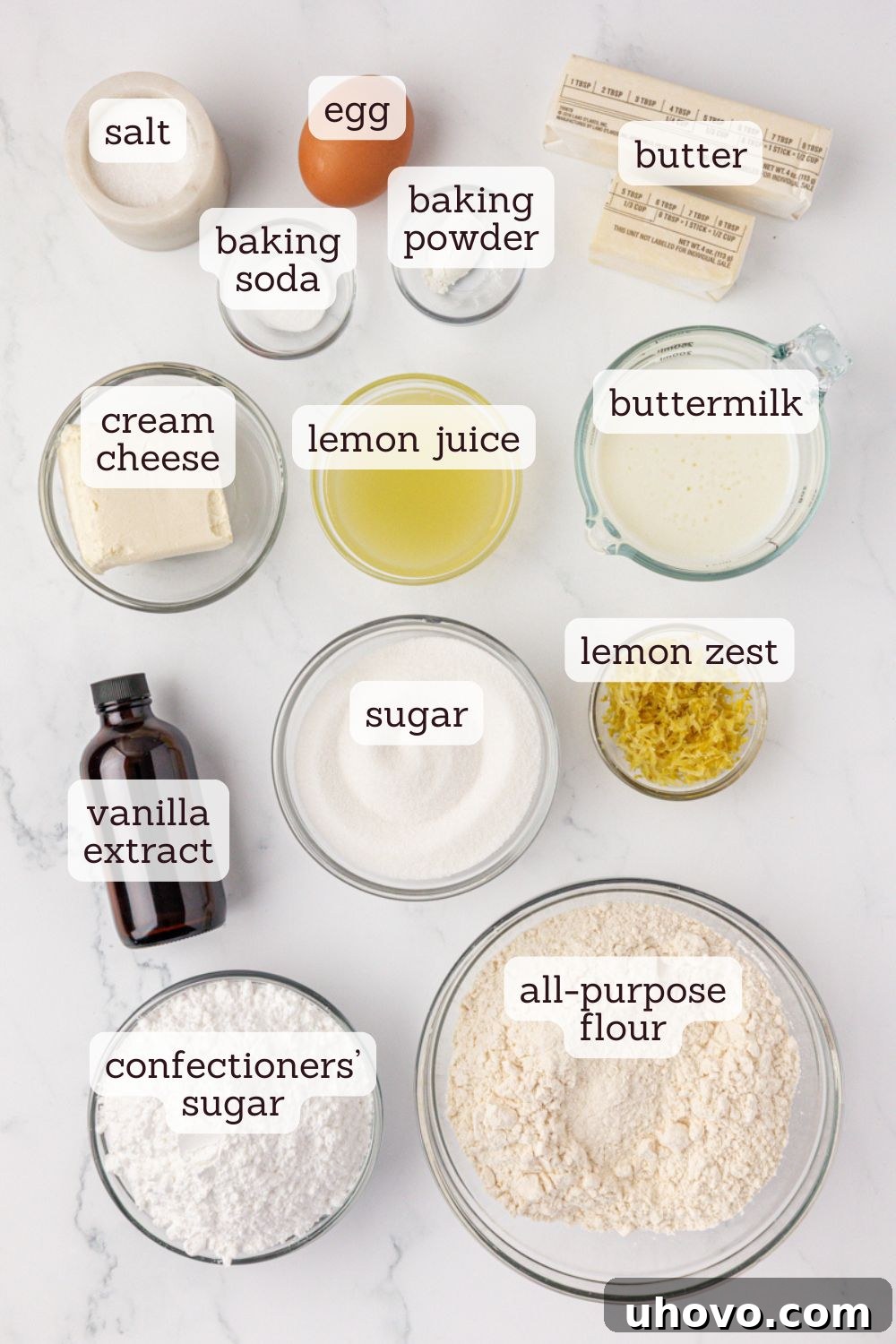 Overhead view of key ingredients for lemon whoopie pies laid out on a white surface, including lemons, butter, cream cheese, sugar, and flour.
